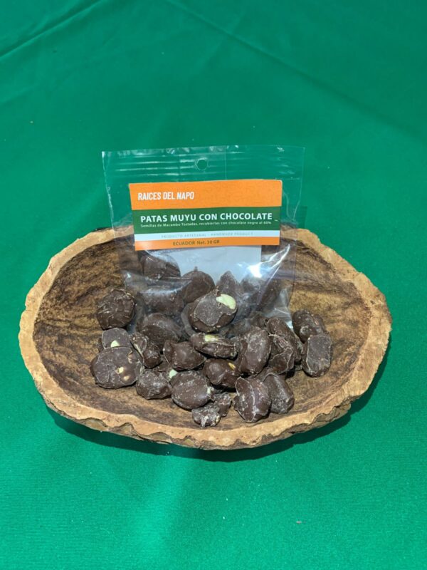 Bolsa de "Patas Muyu con Chocolate" de la marca Raíces del Napo, un snack artesanal de semillas de macambo tostadas cubiertas con chocolate negro, presentado en un recipiente rústico sobre un fondo verde.