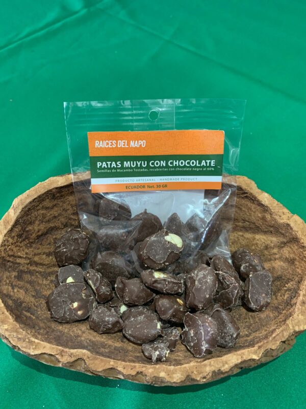 Bolsa de "Patas Muyu con Chocolate" de la marca Raíces del Napo, un snack artesanal de semillas de macambo tostadas cubiertas con chocolate negro, presentado en un recipiente rústico sobre un fondo verde.