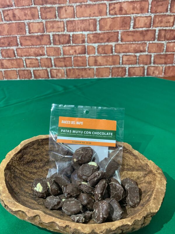 Bolsa de "Patas Muyu con Chocolate" de la marca Raíces del Napo, un snack artesanal de semillas de macambo tostadas cubiertas con chocolate negro, presentado en un recipiente rústico sobre un fondo verde.